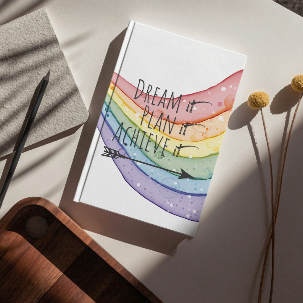 Dream It, Plan It, Achieve It personal development journal displayed on a desk featuring a rainbow pathway cover design for goal setting, planning, self-discovery, and personal growth.